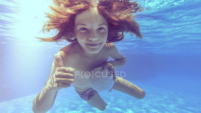 Sonriente niño bajo el agua en una piscina sosteniendo una pelota y haciendo un gesto hacia arriba pulgares - foto de stock