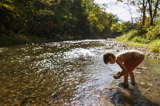 Garçon debout dans une rivière ramassant des roches — Photo de stock