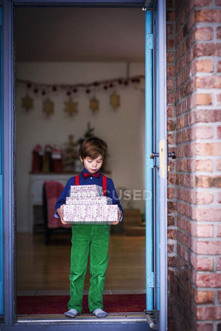 Junge steht in einer Tür und hält einen Stapel verpackter Weihnachtsgeschenke in der Hand — Stockfoto