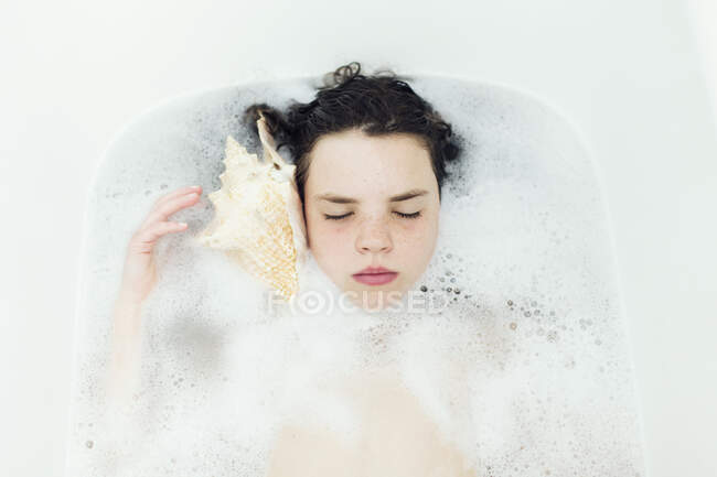 Дівчина лежить у бульбашковій ванні, слухаючи мушлю — стокове фото