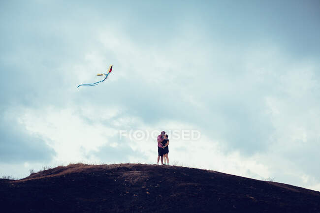 Casal de pé em uma colina voando um papagaio e beijando — Fotografia de Stock