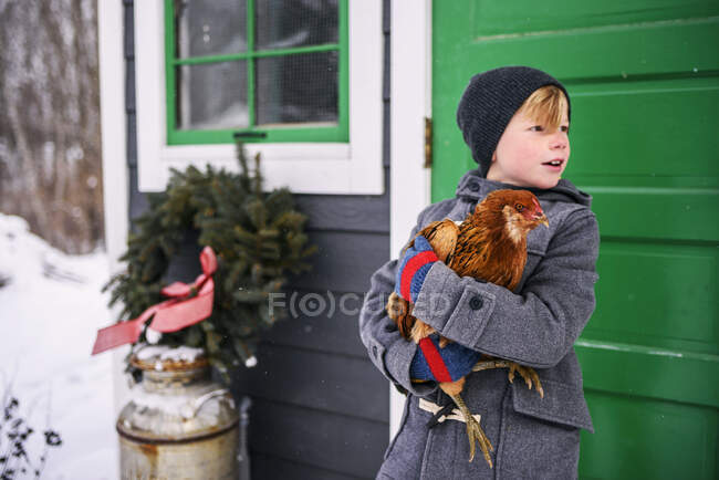 Chico parado afuera de una casa sosteniendo un pollo - foto de stock