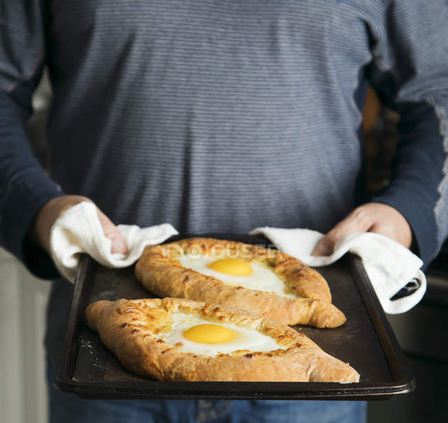 Hombre sosteniendo una bandeja para hornear con dos huevos y queso khachapuri - foto de stock