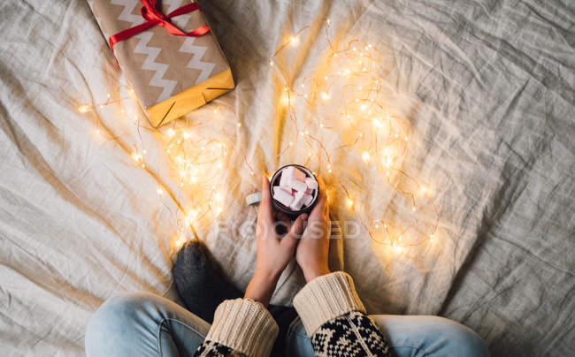 Frau sitzt auf einem Bett und hält einen Becher heiße Schokolade mit Marshmallows in der Hand — Stockfoto