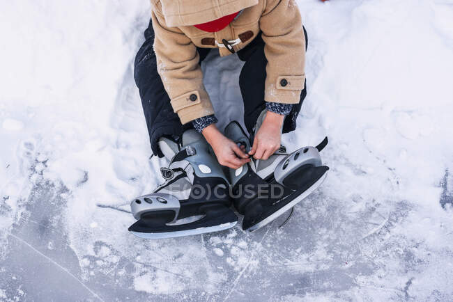 Menino sentado em um lago congelado colocando em seus patins no gelo — Fotografia de Stock