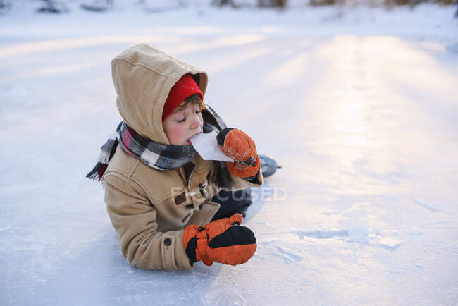 Menino deitado em um lago congelado usando patins de gelo comendo gelo — Fotografia de Stock