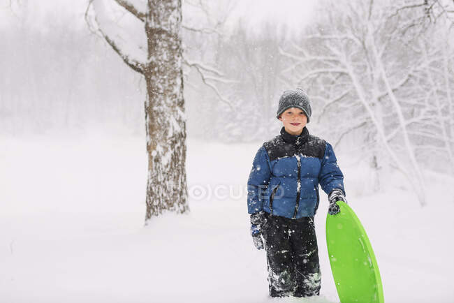 Niño de pie con un trineo en la nieve pesada - foto de stock
