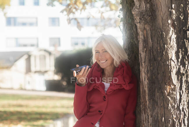 Усміхнена жінка, що схилилася до дерева, розмовляючи на мобільному телефоні (Німеччина). — стокове фото