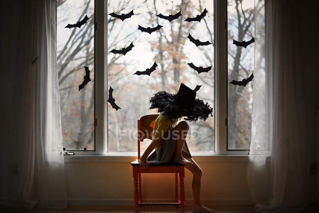 Girl wearing a witches hat sitting on a chair by a window decorated with bats, United States — Stock Photo