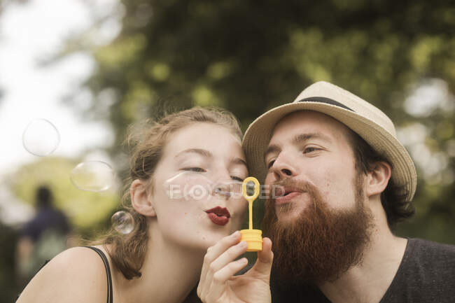 Casal sentado em um parque segurando uma varinha de bolha soprando bolhas — Fotografia de Stock