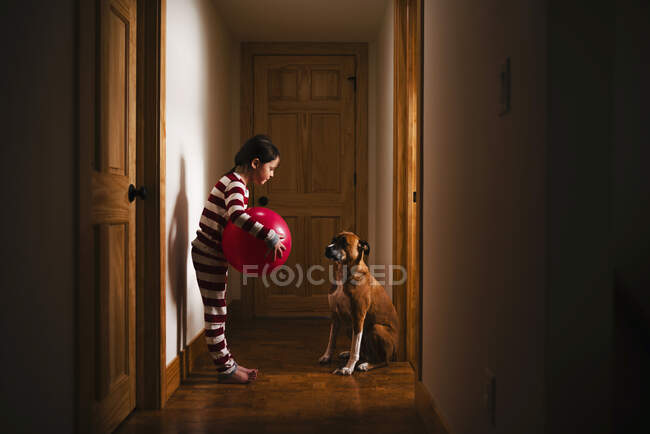 Chica de pie en el pasillo sosteniendo una pelota gigante jugando con su perro - foto de stock