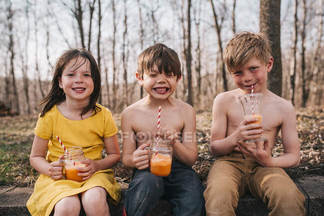 Tres niños sentados en una pared disfrutando de una bebida de verano - foto de stock
