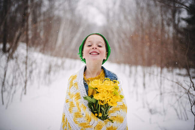 Smiling girl wrapped in a blanket standing in the snow holding a bunch of yellow flowers — Stock Photo