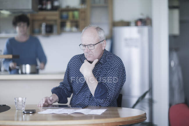 Homem sentado à mesa trabalhando enquanto sua esposa prepara comida — Fotografia de Stock