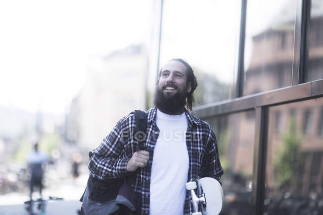 Sorrindo homem andando pela rua carregando um skate — Fotografia de Stock