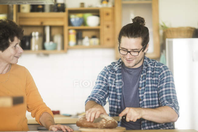 Mère et fils adulte debout dans la cuisine tranchant une miche de pain — Photo de stock