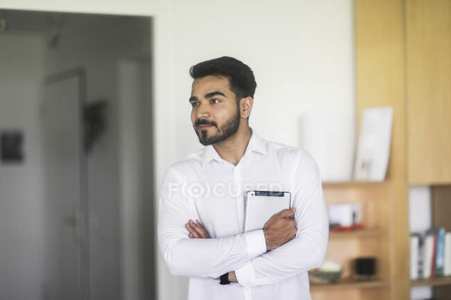 Усміхнений чоловік стоїть у вітальні зі складеними руками, тримаючи цифровий планшет — стокове фото