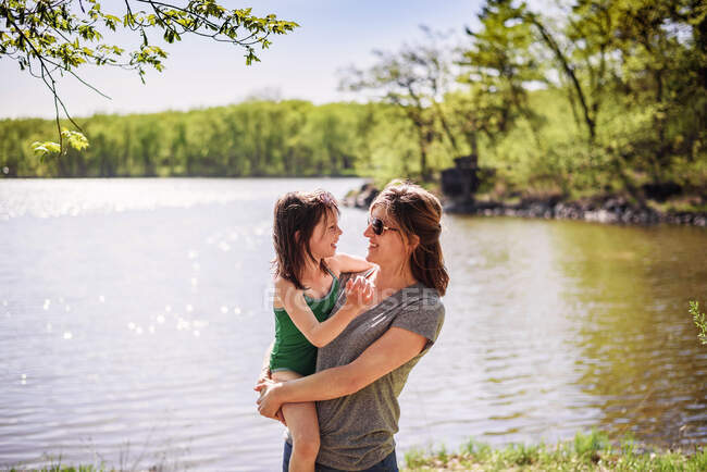 Madre de pie junto a un lago llevando a su hija - foto de stock