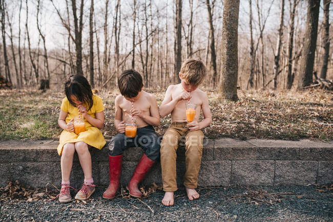Três crianças sentadas em uma parede desfrutando de uma bebida de verão — Fotografia de Stock