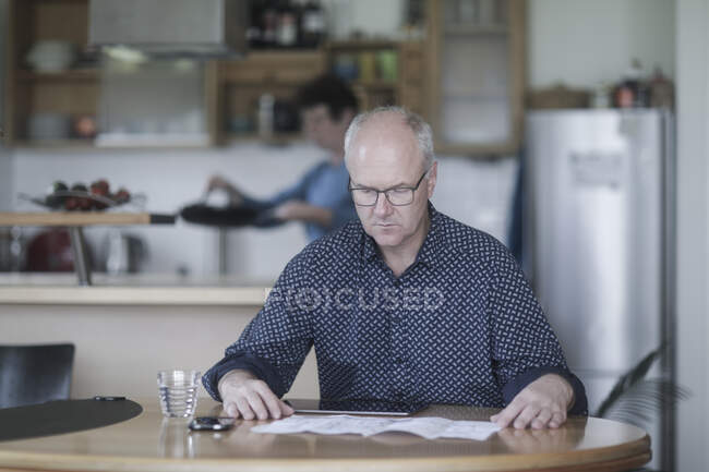 Homem sentado à mesa trabalhando enquanto sua esposa prepara comida — Fotografia de Stock