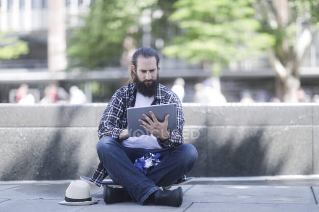 Hombre sentado en su monopatín usando una tableta digital - foto de stock