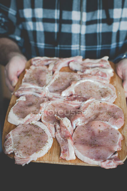Человек держит доску с сырыми приправленными стейками свинины — стоковое фото