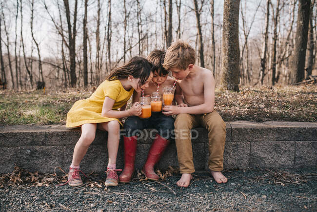 Três crianças sentadas em uma parede desfrutando de bebidas de verão — Fotografia de Stock