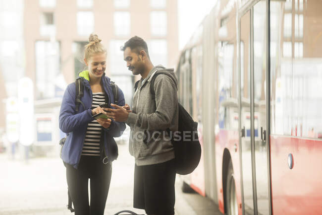 Smiling couple standing by a bus using their mobile phones to plan a journey — Stock Photo