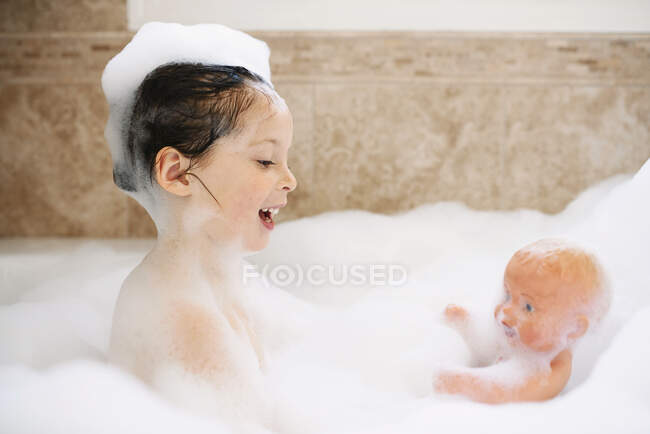 Chica sentada en un baño de burbujas jugando con su muñeca - foto de stock