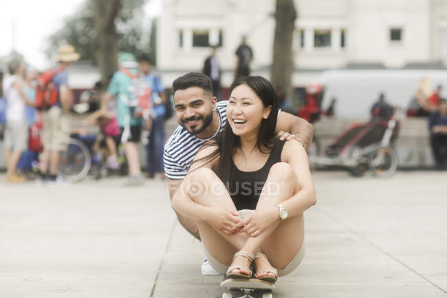 Coppia sorridente seduta su uno skateboard in una piazza della città — Foto stock