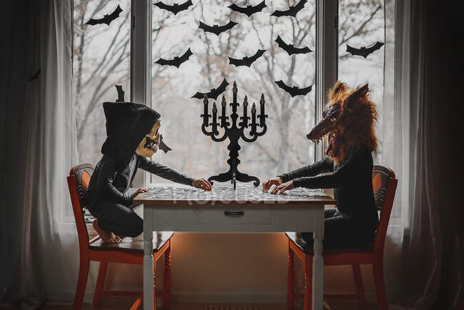 Dos niños con disfraces de Halloween sentados junto a una ventana haciendo un rompecabezas, Estados Unidos - foto de stock