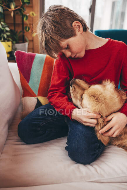 Chico abrazando su golden retriever perro en sofá - foto de stock