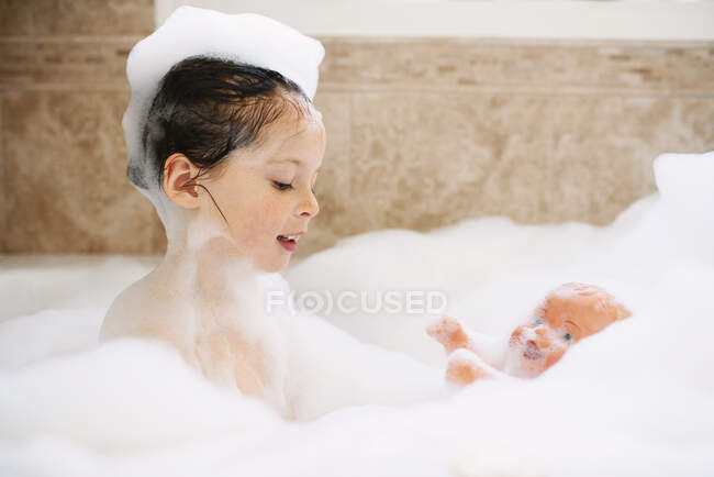 Девушка, сидящая в пенной ванне и играющая со своей куклой — стоковое фото