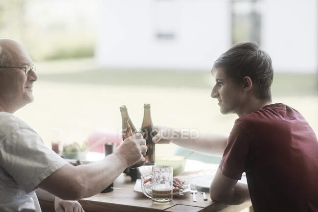 Padre e hijo adultos sentados en la terraza haciendo un brindis de celebración - foto de stock