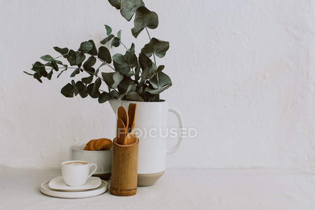 Tasse à café, croissant, ustensiles de cuisine et eucalyptus dans une cruche — Photo de stock