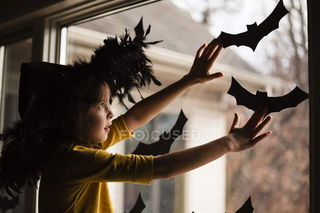 Fille portant un chapeau de sorcières collant des décorations de chauve-souris sur une fenêtre, États-Unis — Photo de stock