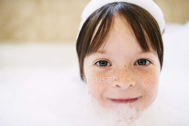 Retrato de uma menina sorridente sentada em um banho de espuma com sabão na cabeça — Fotografia de Stock
