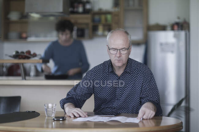 Homme assis à table travaillant pendant que sa femme prépare à manger — Photo de stock