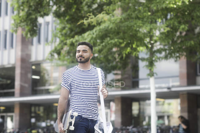 Sorrindo homem andando pela rua carregando um skate — Fotografia de Stock