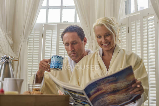 Pareja sentada en una habitación de hotel leyendo una revista y disfrutando del desayuno - foto de stock