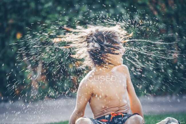 Niño sentado en la hierba sacudiendo su cabello mojado - foto de stock