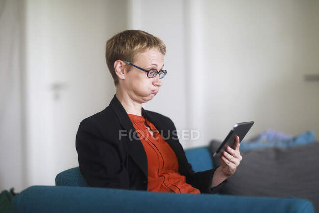 Женщина, сидящая на диване с цифровой табличкой и дующая в щеки — стоковое фото