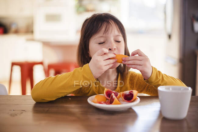 Девушка сидит за столом и ест кровавый апельсин — стоковое фото