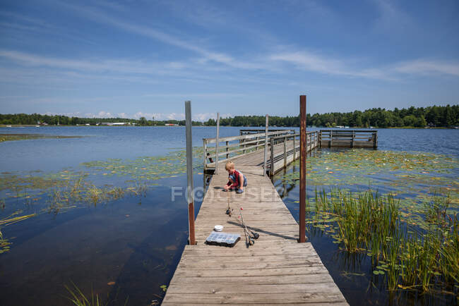 Garçon assis sur un quai attachant appât à sa canne à pêche, États-Unis — Photo de stock