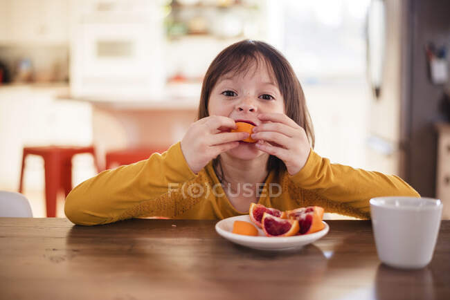 Chica sentada en una mesa comiendo una naranja sangre - foto de stock