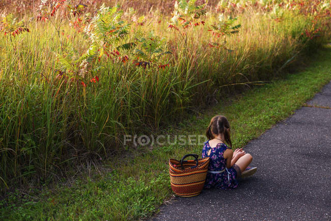 Menina sentada de pernas cruzadas por um campo com uma cesta, Estados Unidos — Fotografia de Stock