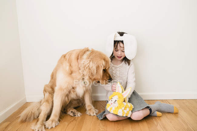 Retrato de una chica sonriente con orejas de conejo sentada en el suelo con su perro - foto de stock
