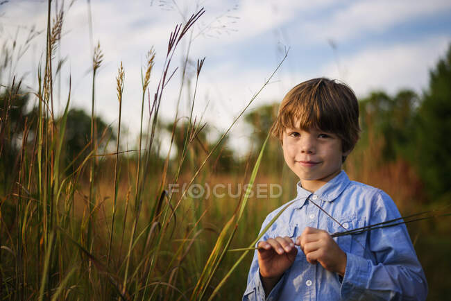 Portrait d'un garçon souriant debout dans un champ au coucher du soleil cueillant de l'herbe longue, États-Unis — Photo de stock