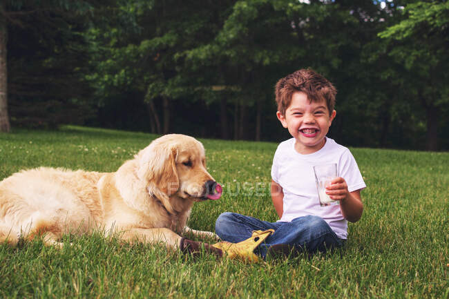 Niño sentado al aire libre bebiendo un vaso de leche con un golden retriever - foto de stock