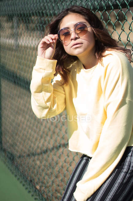 Teenage girl adjusting her sunglasses, leaning against a mesh fence — Stock Photo